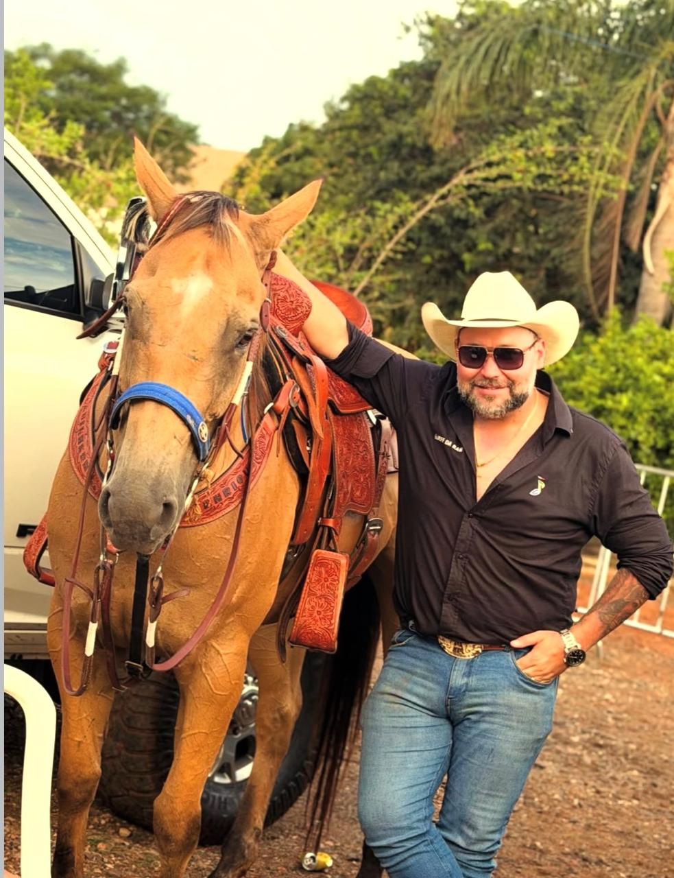 Jeff da RAM apoiado na sela de um cavalo baio, ao lado de uma RAM branca, no fim da tarde